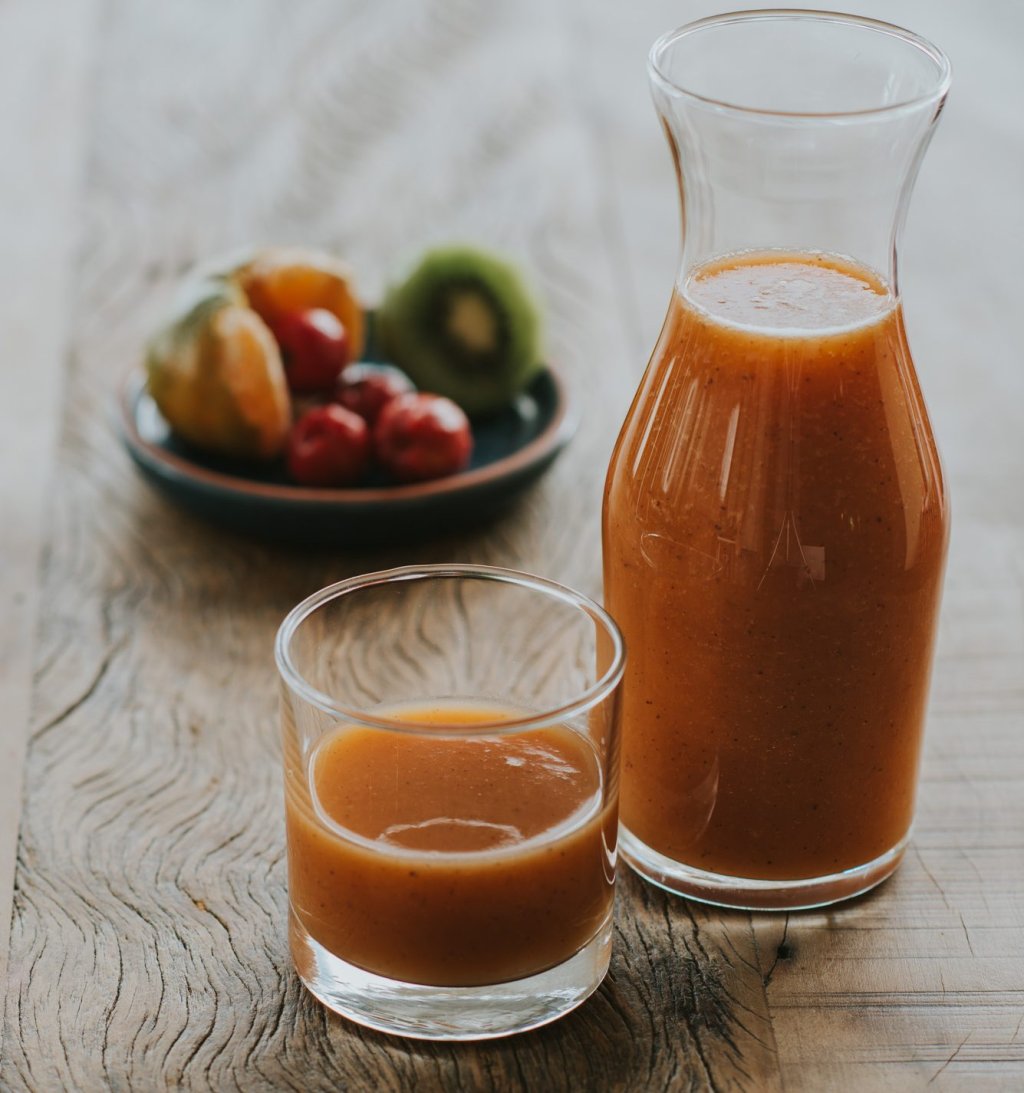 copo de suco de cor laranja e jarra com suco da mesma cor aparecem no primeiro plano. Ao fundo, desfocado, uma cesta com frutas cítricas