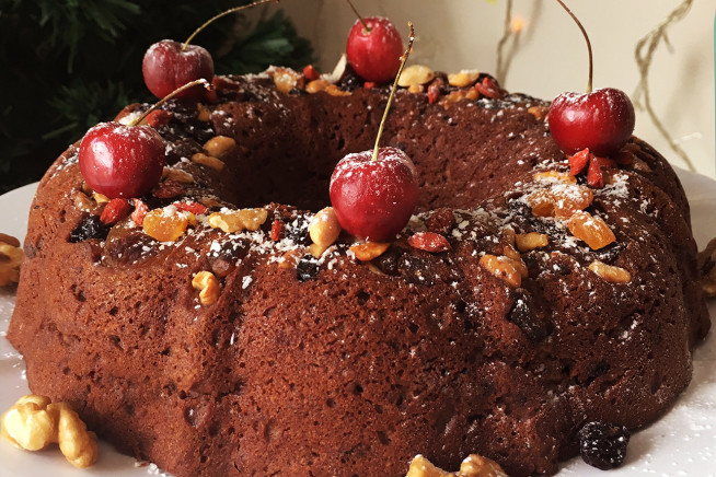 A imagem contém um bolo redondo com furo no meio de cor marrom coberto por frutas cristalizadas, cerejas e polvilhado com açúcar