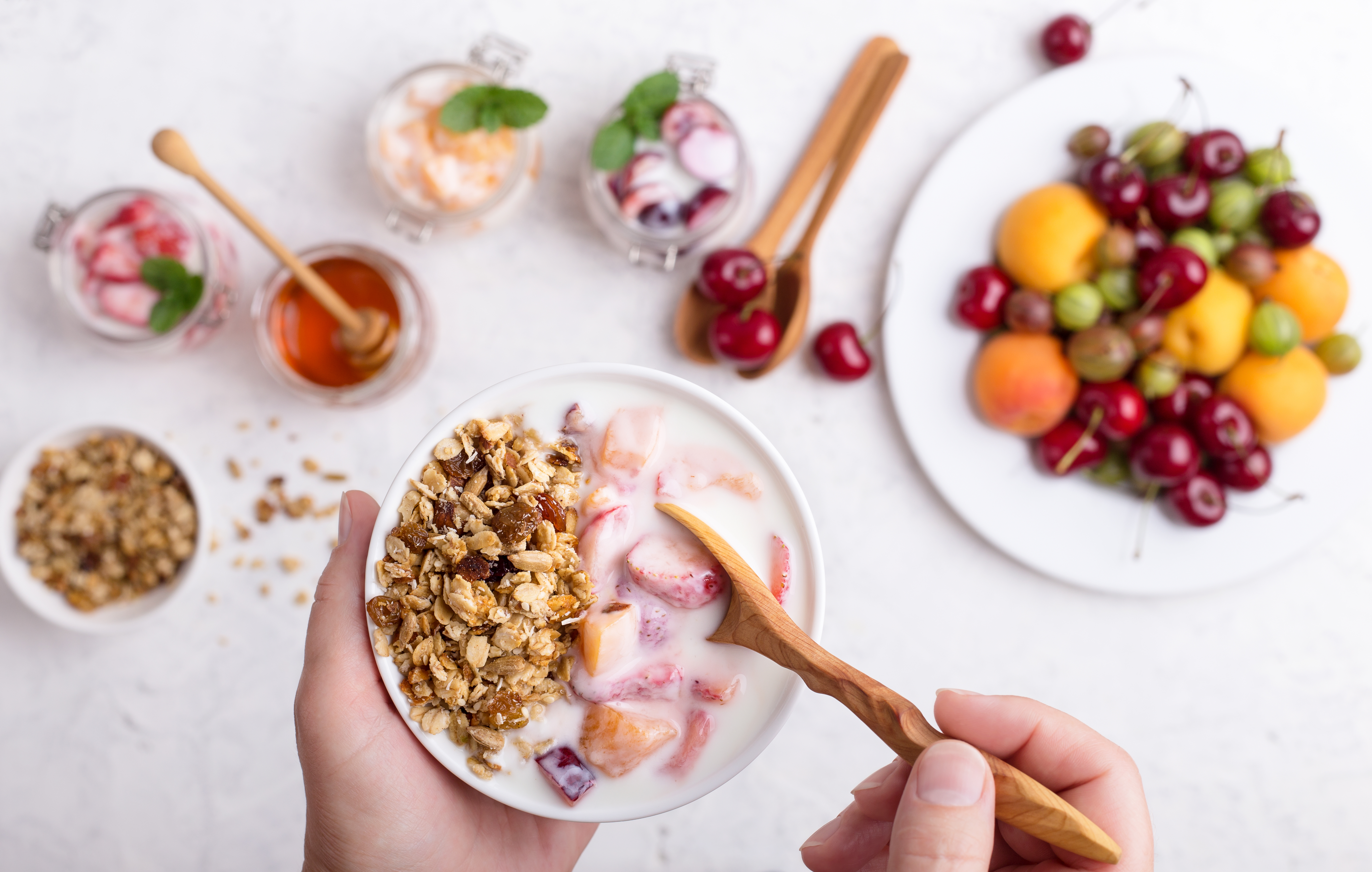 Cropped hands of woman holding bowl of cereals. Healthy breakfast, favorite meal. Homemade granola, fresh summer fruits and berries, yoghurt with sweet cherry, strawberry and apricot on light gray table viewed from above