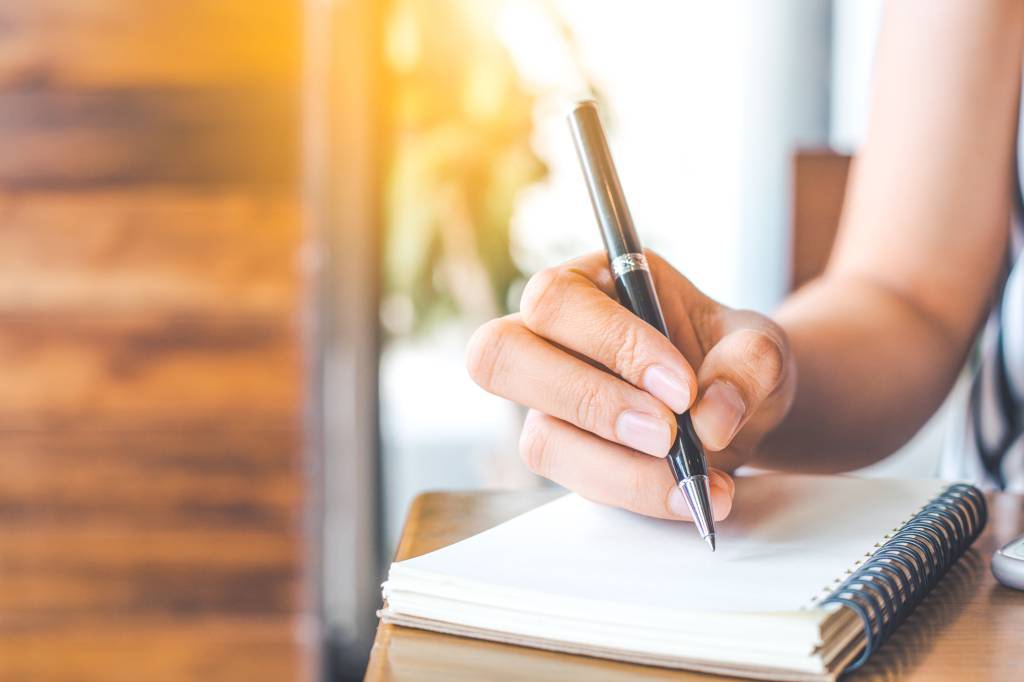woman’s hand is writing on a blank notepad with a pen on a wooden desk.