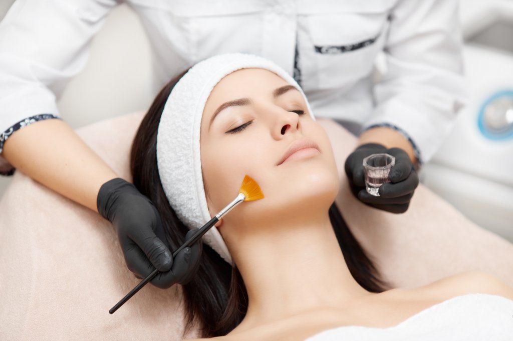 Beautiful woman with smooth skin relaxing and enjoying spa procedures. Beautician applying anti-acne mask, making face peeling. Concept of facial, rejuvenation and regeneration of skin.