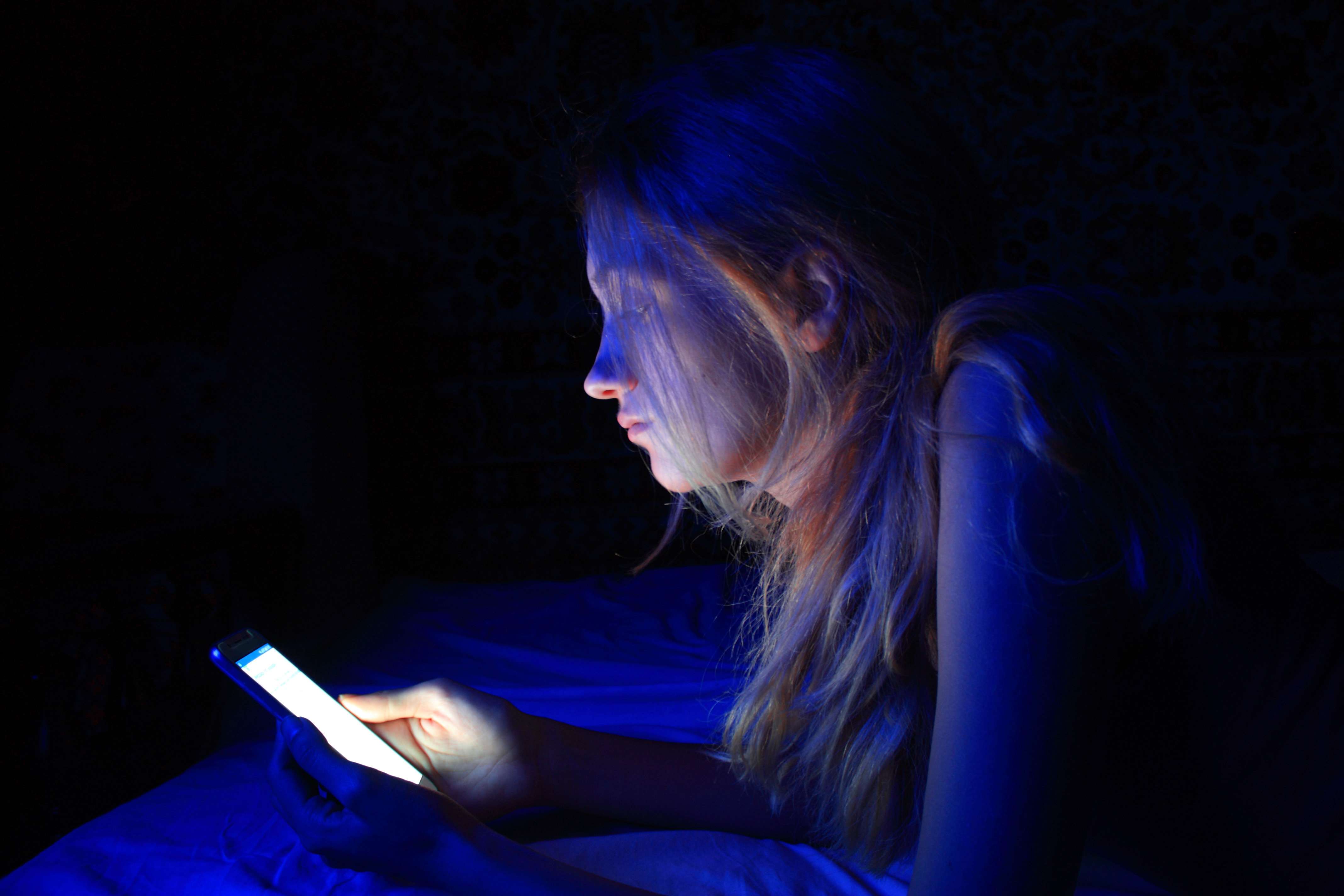 Mulher deitada na cama mexendo no celular que está emitindo uma luz azul