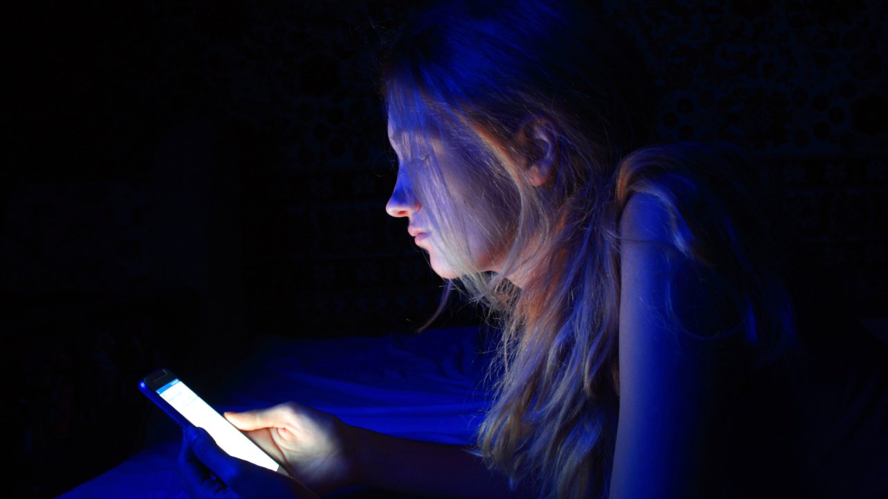Mulher deitada na cama mexendo no celular que está emitindo uma luz azul
