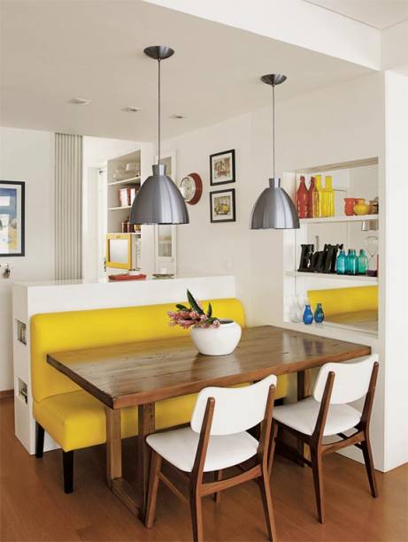 Na área de refeições, um sofá amarelo de couro sintético é o destaque da decoração. Uma mesa de cadeira maciça é servida por cadeiras, que acrescenta branco à combinação quente. Uma estante embutida, com fundo espelhado, amplia visualmente este canto. Projeto <span>das arquitetas  Isabella Lucena e Joanna Fraga.</span>