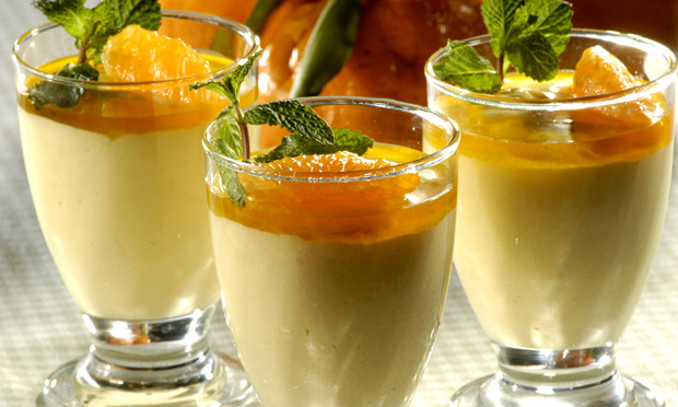 Três taças de sobremesa com musse com amarelo pastel coberto por calda de cor laranja e decorado com folhinhas verdes. Ao fundo, desfocada, aparece uma tangerina