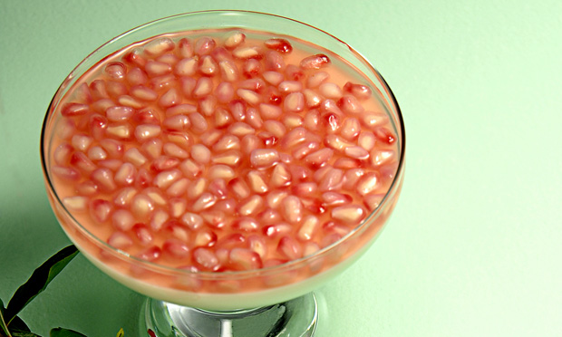 Em uma taça de vidro aparecem muitas sementes de romã envolvidas por um líquido avermelhado e translúcido. A taça está sobre uma superfície verde
