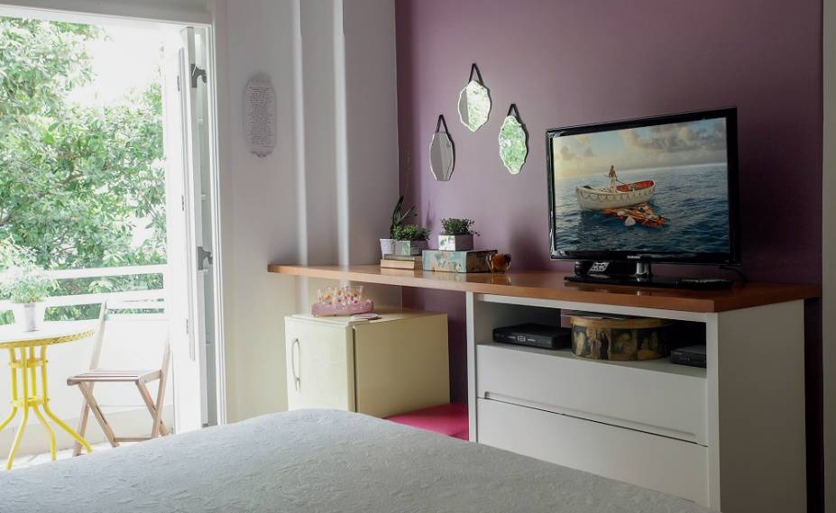 <span>Na frente da cama, a parede da TV se enfeitou com um tom rosado e um kit de espelhos decorativos. A bancada que sustenta o aparelho é um tampo de MDF, recortado conforme os desníveis da alvenaria vizinha, que se apoia na antiga cômoda e vai aparafusado na extremidade. Dessa forma, criou-se um nicho para  o frigobar retrô, sonho  de consumo do casal. Decorado pela blogueira e decoradora carioca Vivi Visentin, do blog Decorviva.</span>