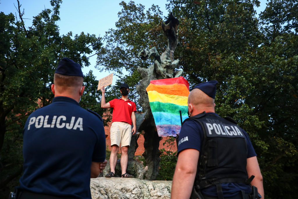 Ativista LGBT segura bandeira do arco-íris, símbolo da comunidade, em uma estátua enquanto segura uma placa com os dizeres "Apoie Margot", ativista que foi presa durante protestos a favor de direitos LGBT, no último sábado (8), na Polônia.
