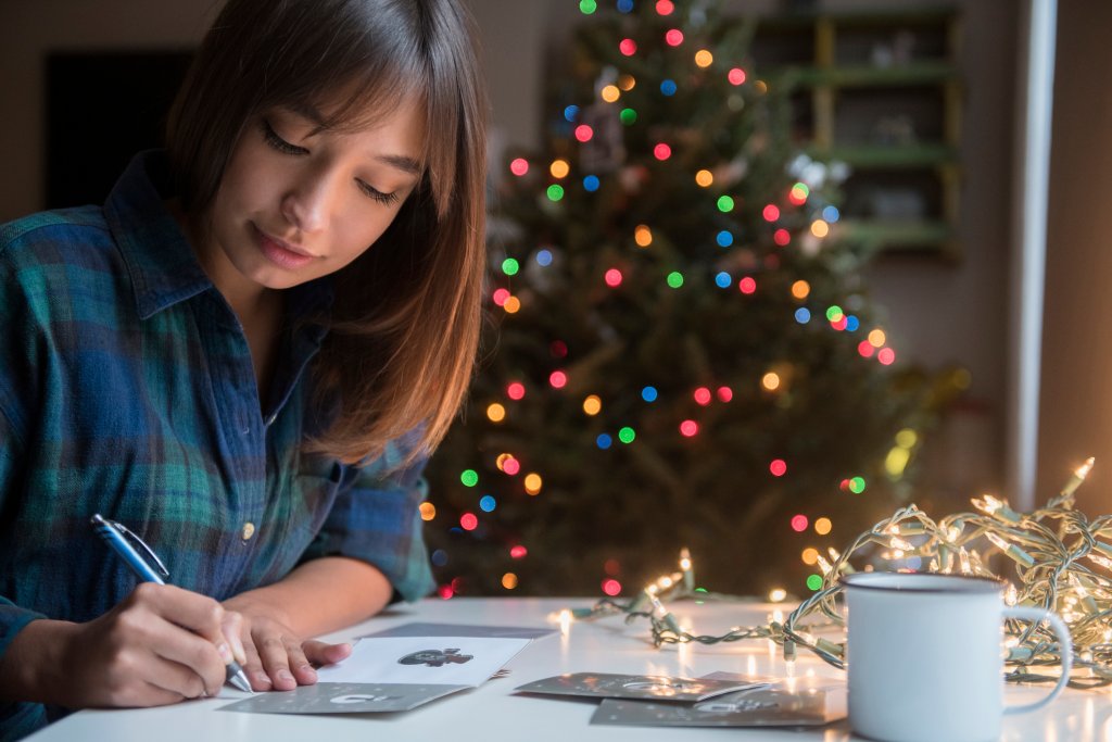 Mulher sentada perto de uma árvore de Natal enquanto escreve uma mensagem em um cartão