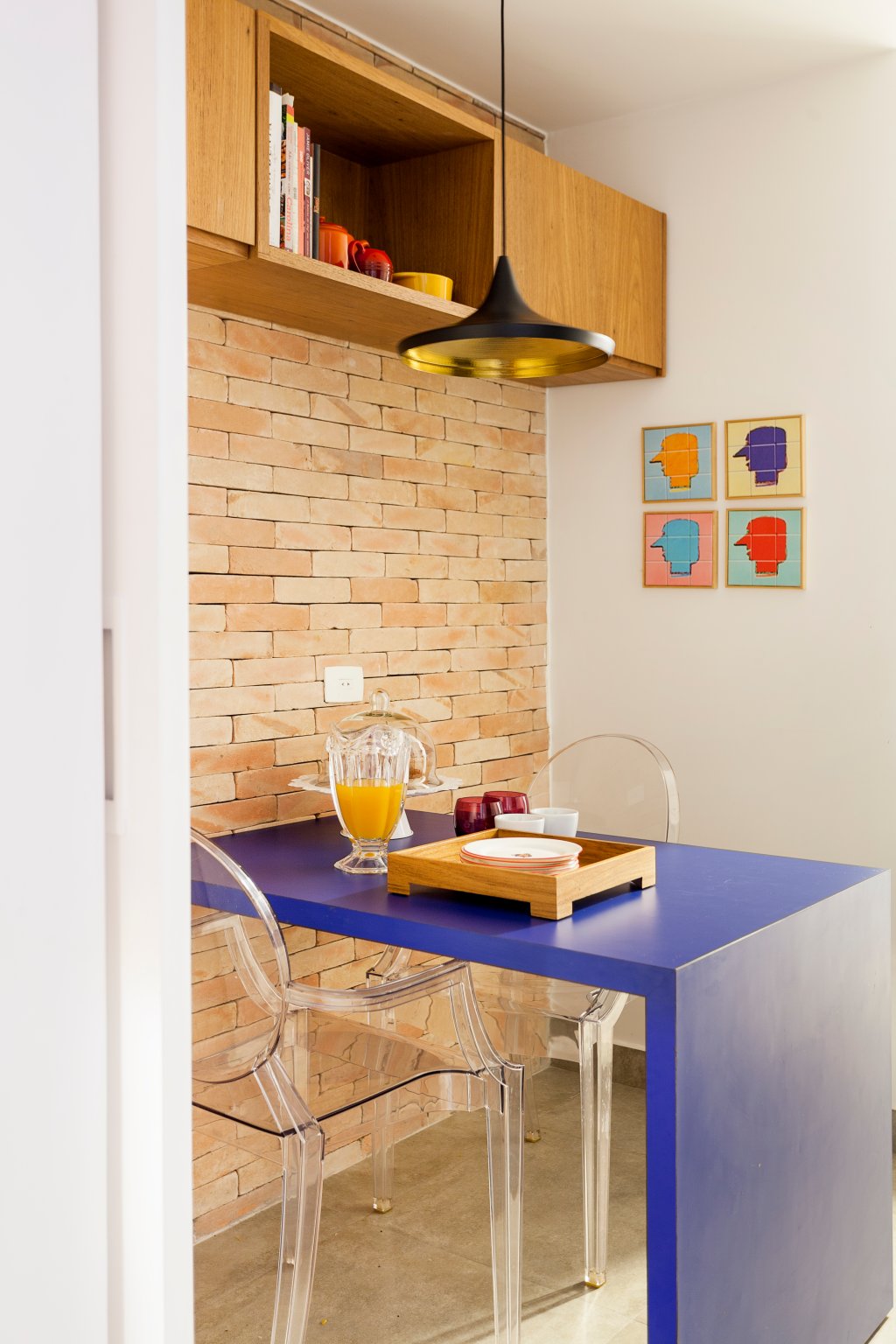 Na foto, em diagonal, é retratada uma cozinha, com mesa azul e cadeiras transparente. A parede ao fundo é de tijolos aparentes. Há um quadro colorido na paredes. O pendente preto e dourado aparece sobre a mesa, que tem uma bandeja de madeira e jarra de vidro com suco de laranja.