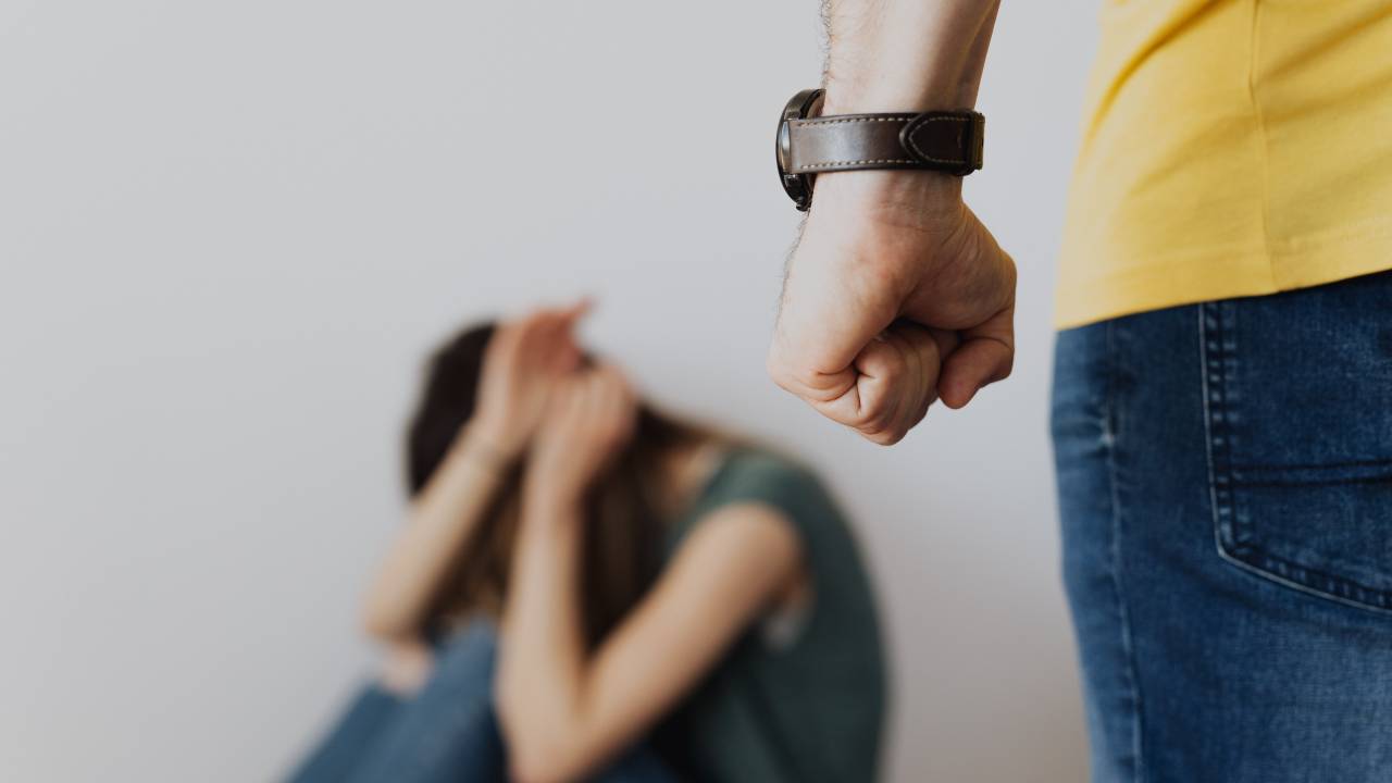 Ao fundo, uma mulher sentada no chão coloca as mãos na frente do rosto para se proteger da possível agressão de um homem que aparece no canto direito da imagem, com o punho cerrado