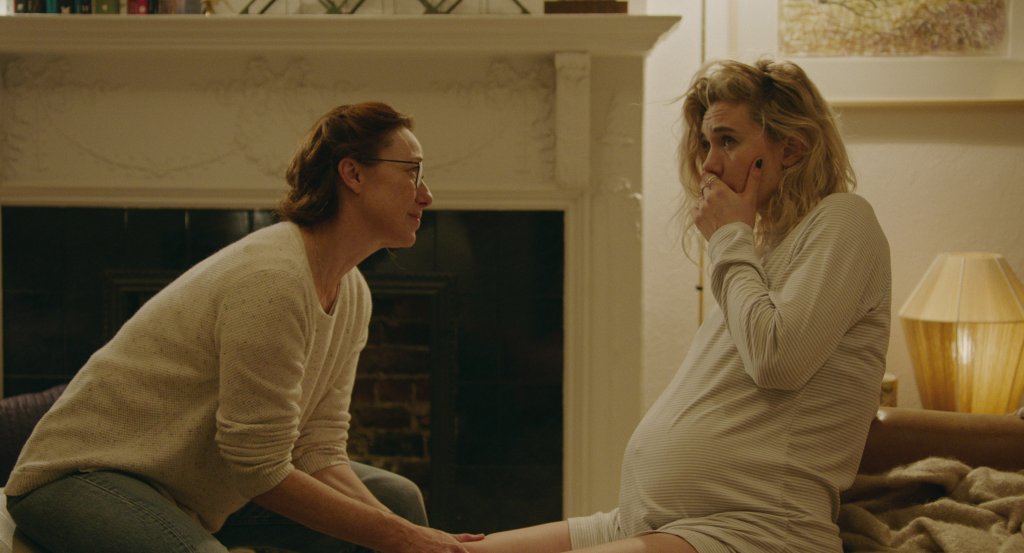 Duas mulheres sentadas olham uma para outra. Uma delas está grávida e com a mão na boca.