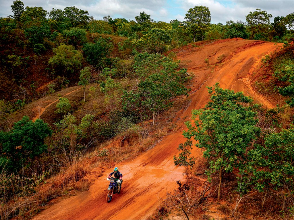 Uma faixa aberta em meio à vegetação vira pista no cenário de terra, com chão vermelho. Uma moto, pilotada por uma mulher, aparece no centro da pista