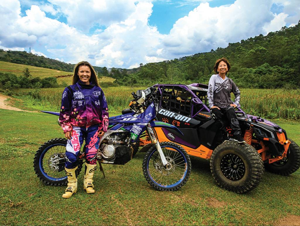 Helena-Deyama-Moara-Sacilotti-rally-dos-sertoes Em um campo verde, duas mulheres vestidas com uniformes estão paradas junto aos seus veículos. Uma delas, loira, tem uma moto, usa uniforme rosa e roxo e botas amarelas. A outra, de cabelos castanhos curtos, usa uniforme cinza e preto e está sentada sobre um UTV