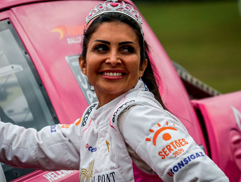 Uma mulher de cabelos escuros presos sorri para a foto. Ela usa um uniforme de corrida e uma coroa. Está apoiada em sua caminhonete pink