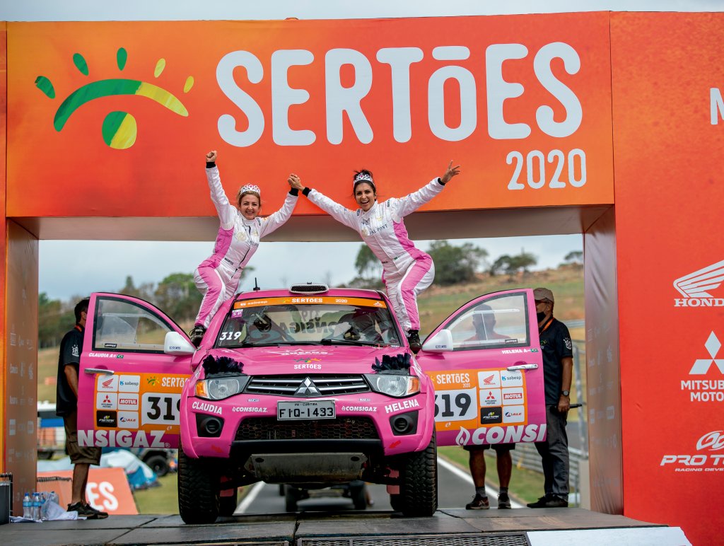 duas mulheres estão de pé apoiadas nas portas abertas de um carro cor de rosa. O farol do carro tem um item que imita cílios