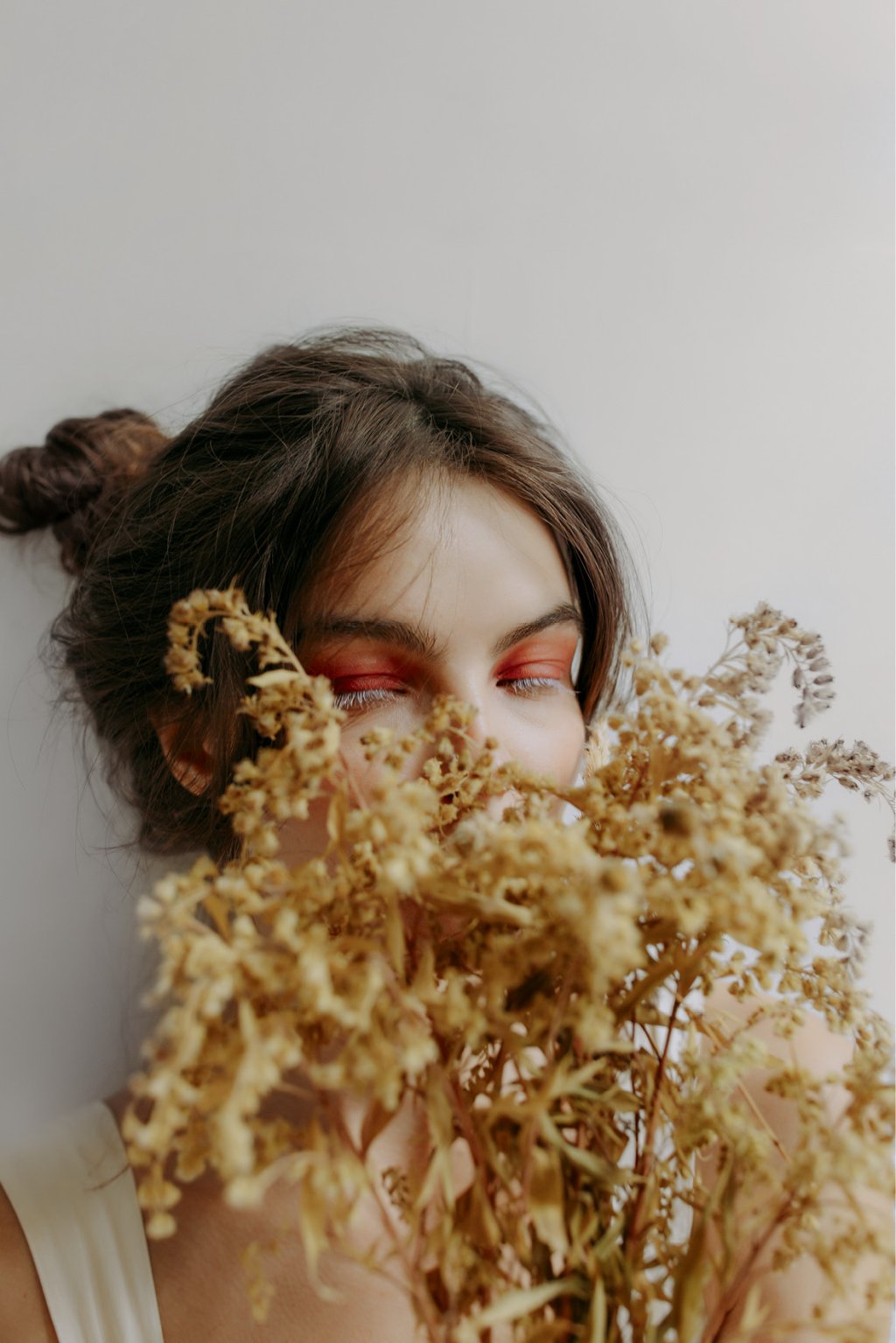 mulher branca com cabelo castanho preso em um coque está maquiada com sombra laranja e rímel branco. Ela segura um buquê de flores secas em frente ao rosto