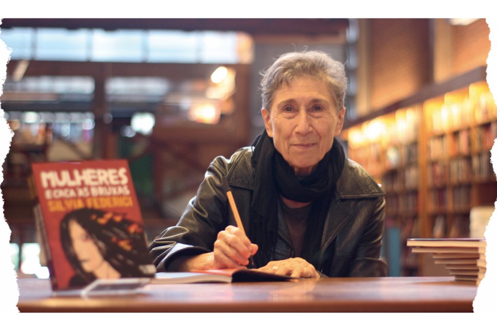 Silvia é uma mulher de cabelos curtos e grisalhos. Ela está sorrindo para a foto num cenário de livraria, enquanto autografa livros