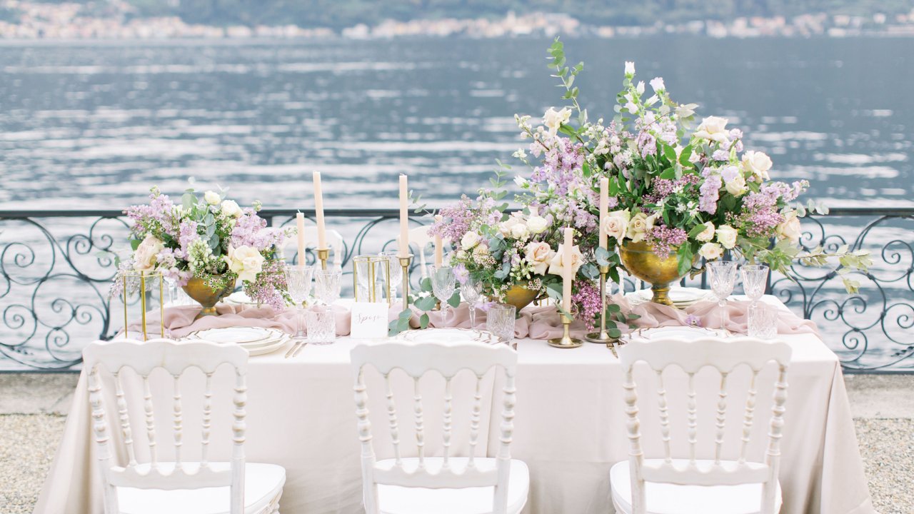 Mesa com toalha branca, e muitas flores está montada para receber convidados em uma festa