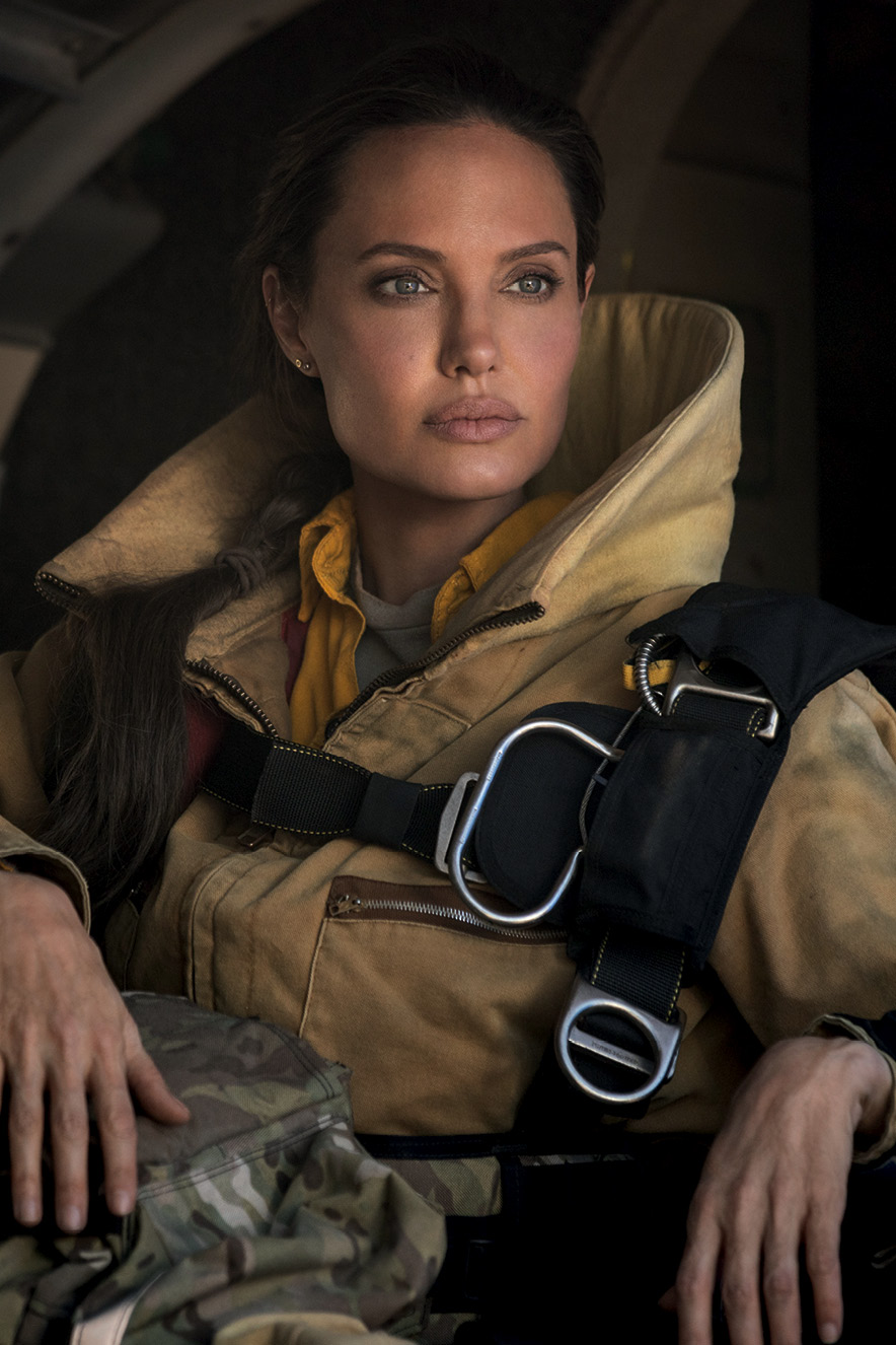Angelina Jolie olha para o horizonte usando um grande casaco e usando cinto de segurança