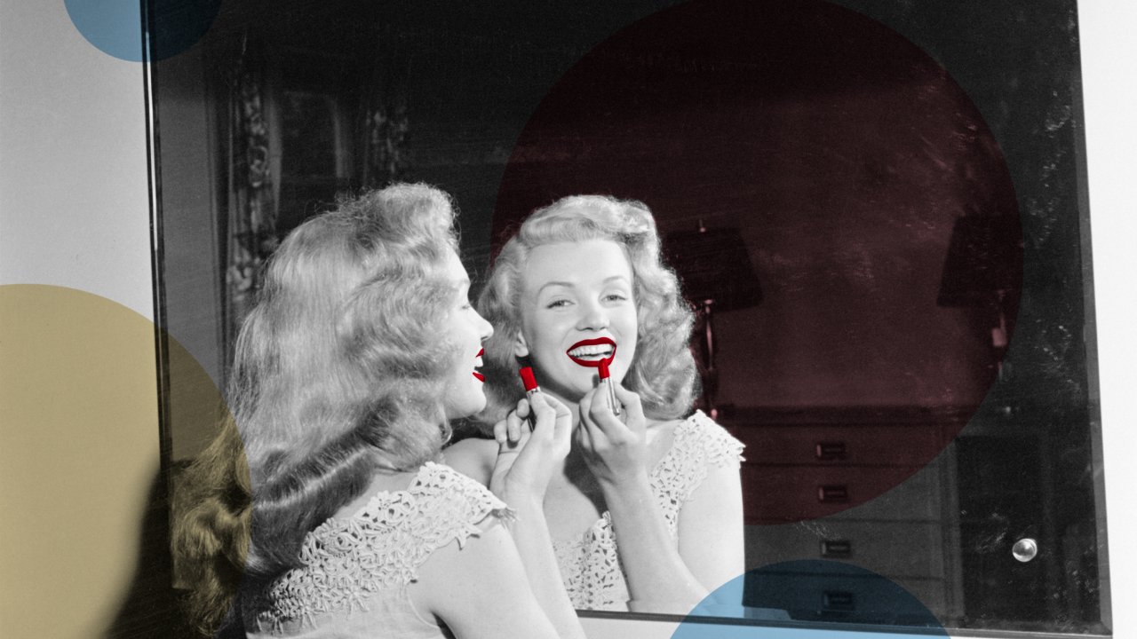 Marilyn Monroe de frente para o espelho passando batom vermelho e sorrindo.