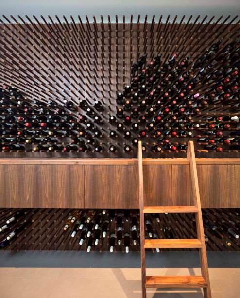 Parecido com um pegboard com diversos suportes, o painel permite acomodar inúmeras garrafas de vinho. O armário, que serve de apoio para a escada, complementa a adega.