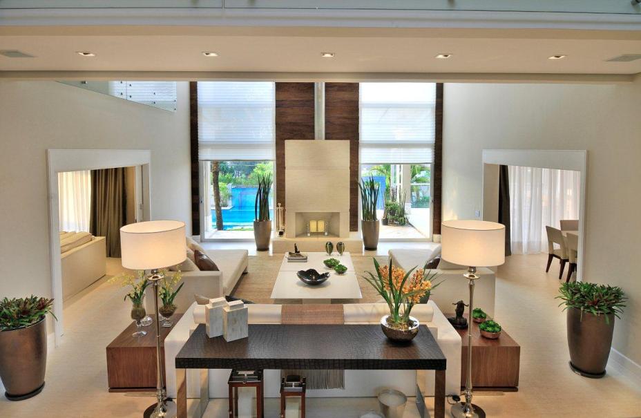 Na sala ampla e com decoração elegante, que mistura off white e madeira escura, a lareira é emoldurada por duas grandes janelas com vista para a piscina. Na sala ampla e com decoração elegante, que mistura off white e madeira escura, a lareira é emoldurada por duas grandes janelas com vista para a piscina.