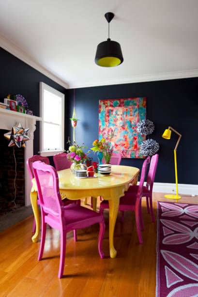 Na sala de jantar, a mesa amarela e as cadeiras pink contrastam com o fundo azul marinho das paredes. Os objetos de decoração que rodeiam o conjunto do jantar também são multicoloridos.