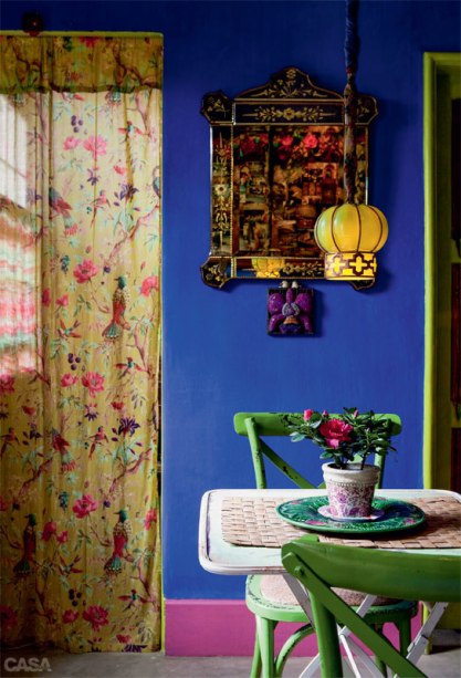 O canto da sala de entrada é uma amostra das cores e estampas que se espalham pela casa da decoradora Silvia Adami. No ambiente, parede azul, rodapé rosa, batentes das portas e cadeiras verdes, além da cortina e da luminária amarelas, se misturam com alegria.