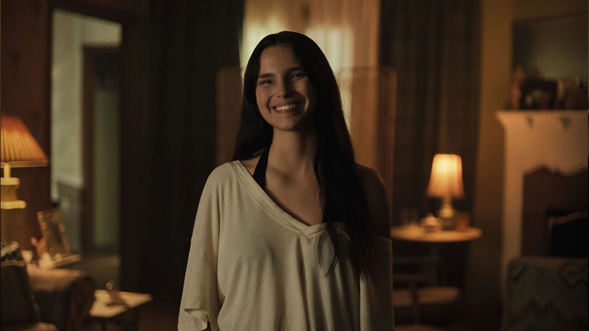 Mulher jovem de cabelos longos e escuros, sorrindo, vestindo camiseta bege de decote V e alça preta, em ambiente interno com iluminação baixa e abajures acesos