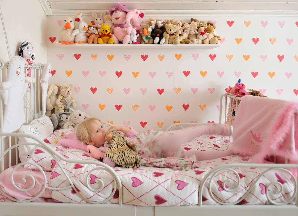 Herzchenmotive auf Bett und Wand im Kinderzimmer; Stofftiersammlung auf Holzbord, darunter blondes Mädchen auf Metallbett