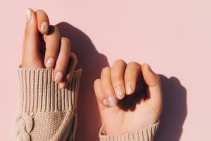 Glowing hands in sweater with knitted sleeves and with nude color manicure with gold particles on pink table. Trendy colors of the year