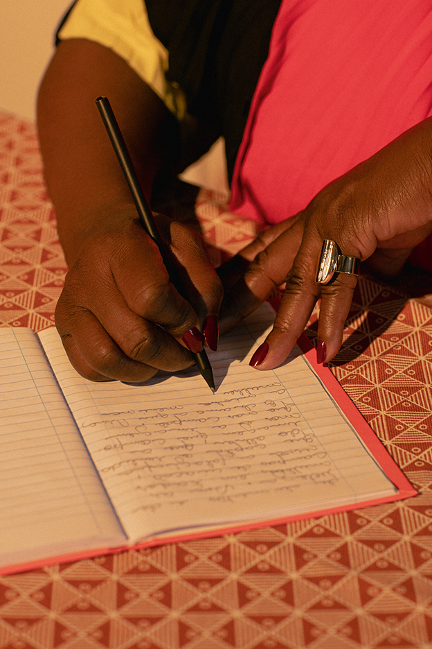 Conceição anota suas inspirações num caderno vermelho. Ela usa vestido do Apartamento 03 e anel, Zâmbia.