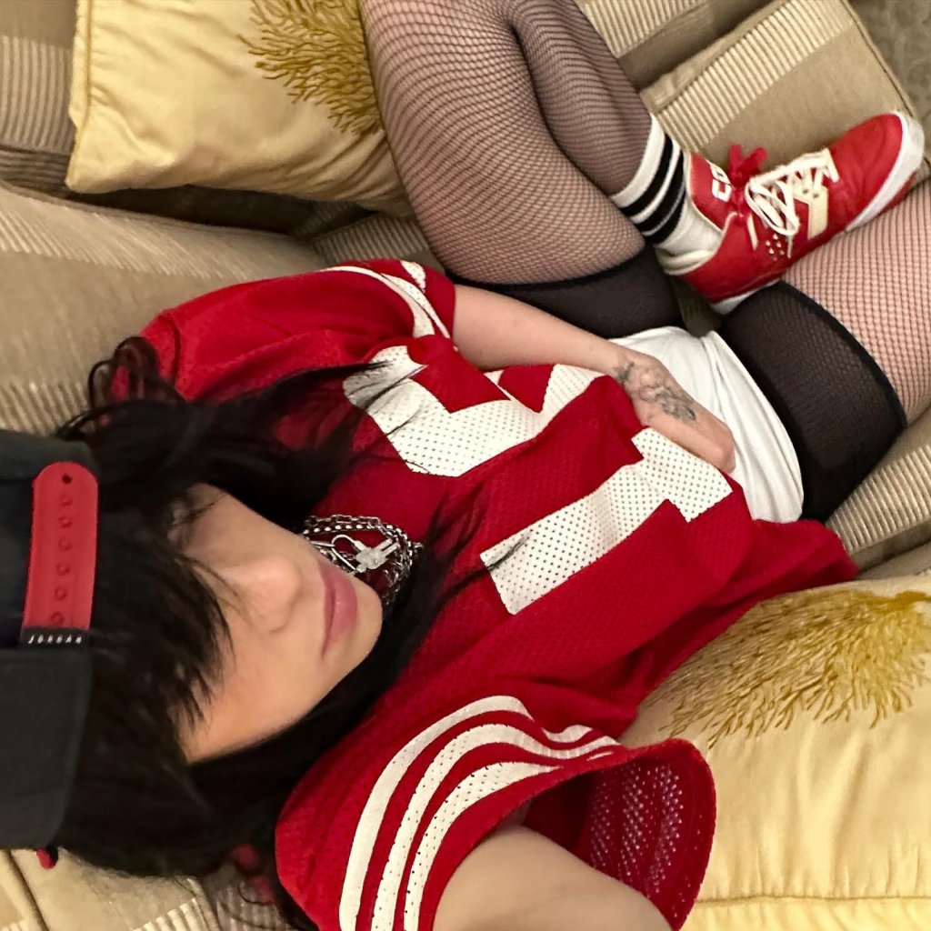 Cantora Billie Eilish sentada em um sofá, vestindo camisa vermelha com sobreposições de meia-calça e shorts preto, característico do Blokecore