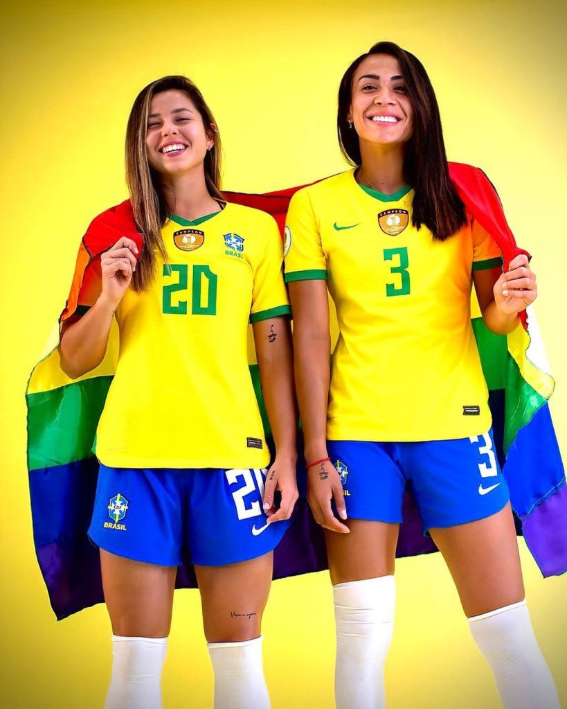 Kathellen e Fernanda Palermo vestidas com o uniforme da seleção e segurando uma bandeira do orgulho LGBTQIAP+