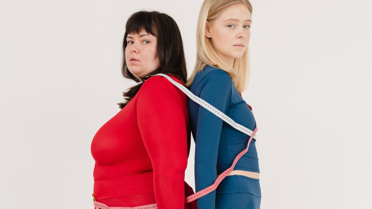 Duas mulheres, uma gorda e uma magra, juntas, com uma fita métrica ao redor do corpo