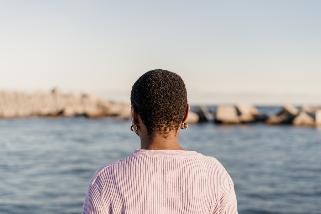 Mulher negra de cabelos raspados de costas, olhando para o mar a sua frente, indicando melancolia