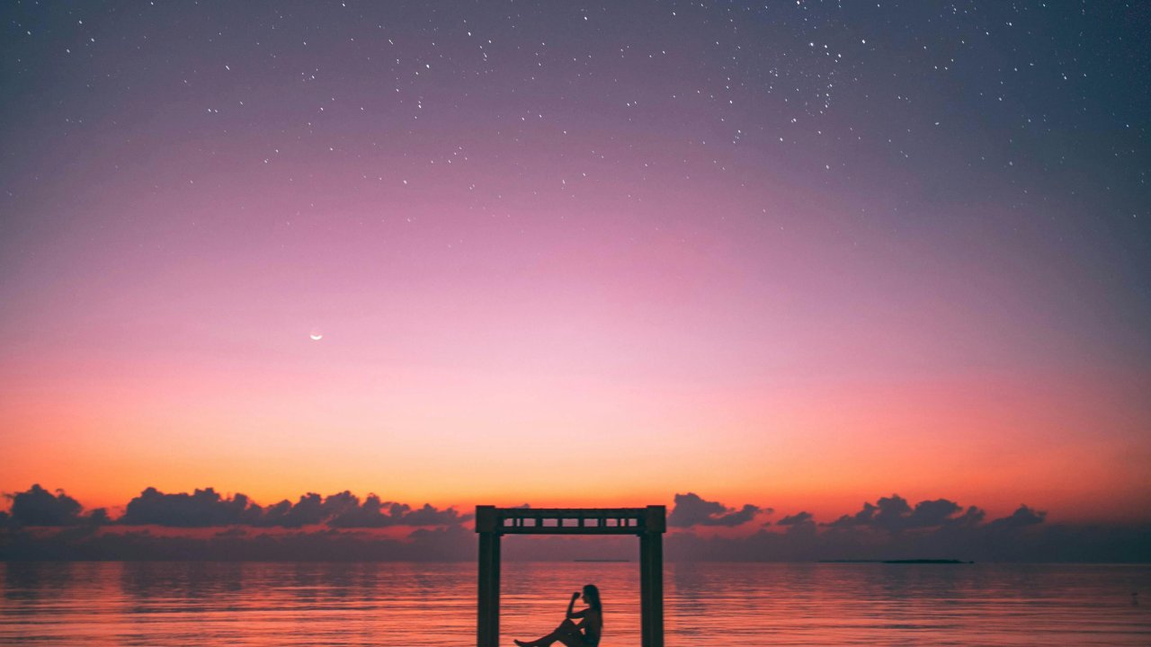 Horóscopo da semana: confira as previsões da semana de 17 a 23 de junho. Imagem Pessoa sentada na beira do rio com um por do sol em tons de rosa e laranja ao fundo, com sentimentos de esperança e confiança nas previsões astrológicas.