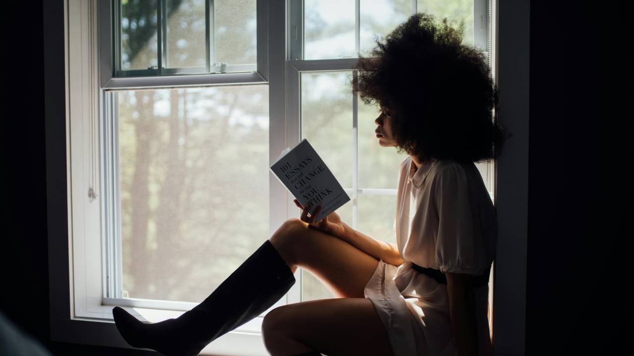 Imagem de uma mulher negra lendo um livro enquanto está sendo em frente a uma janela