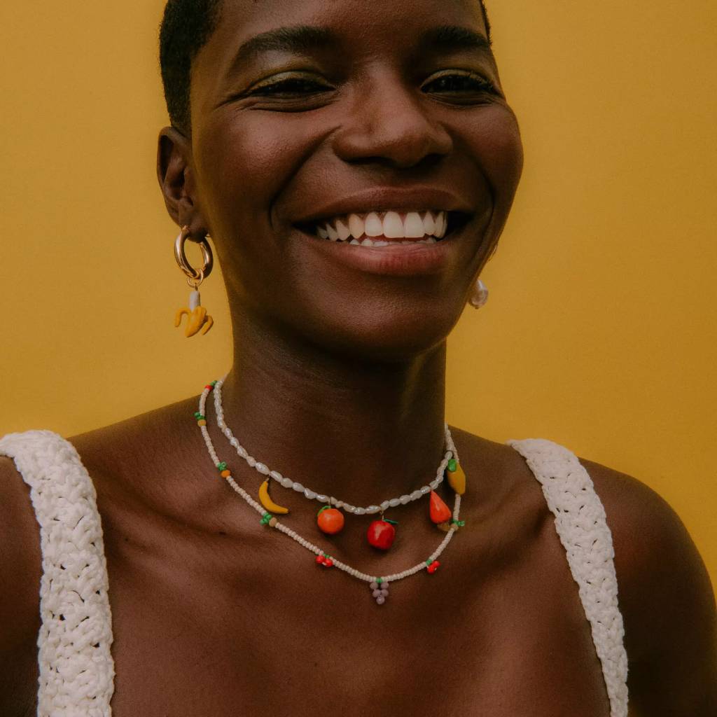 Imagem de mulher sorrindo, vista de frente com um colar branco com de talhes de frutinhas