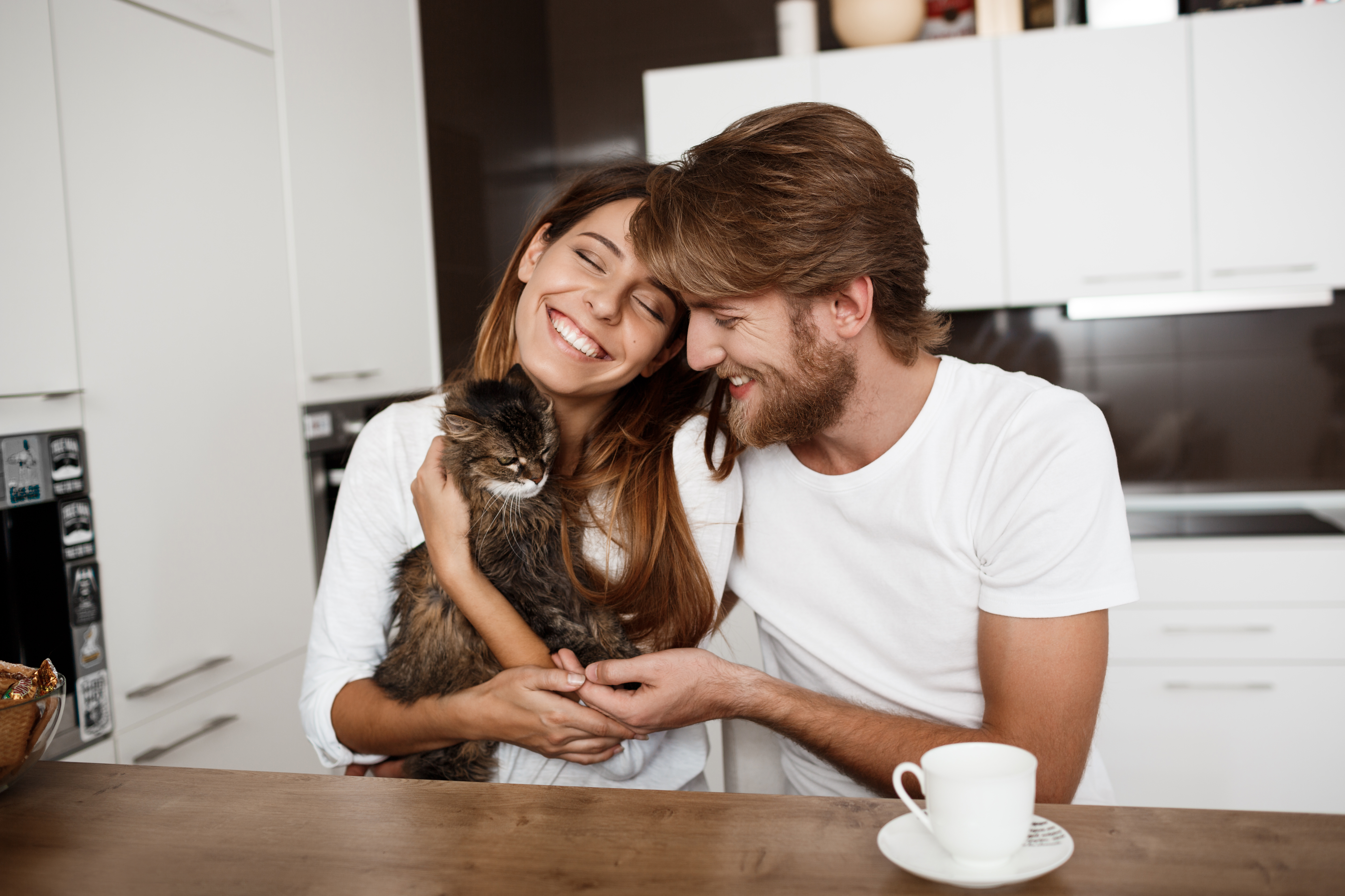 Homem e mulher sentados sorrindo em uma cozinha, a mulher está segurando um gato