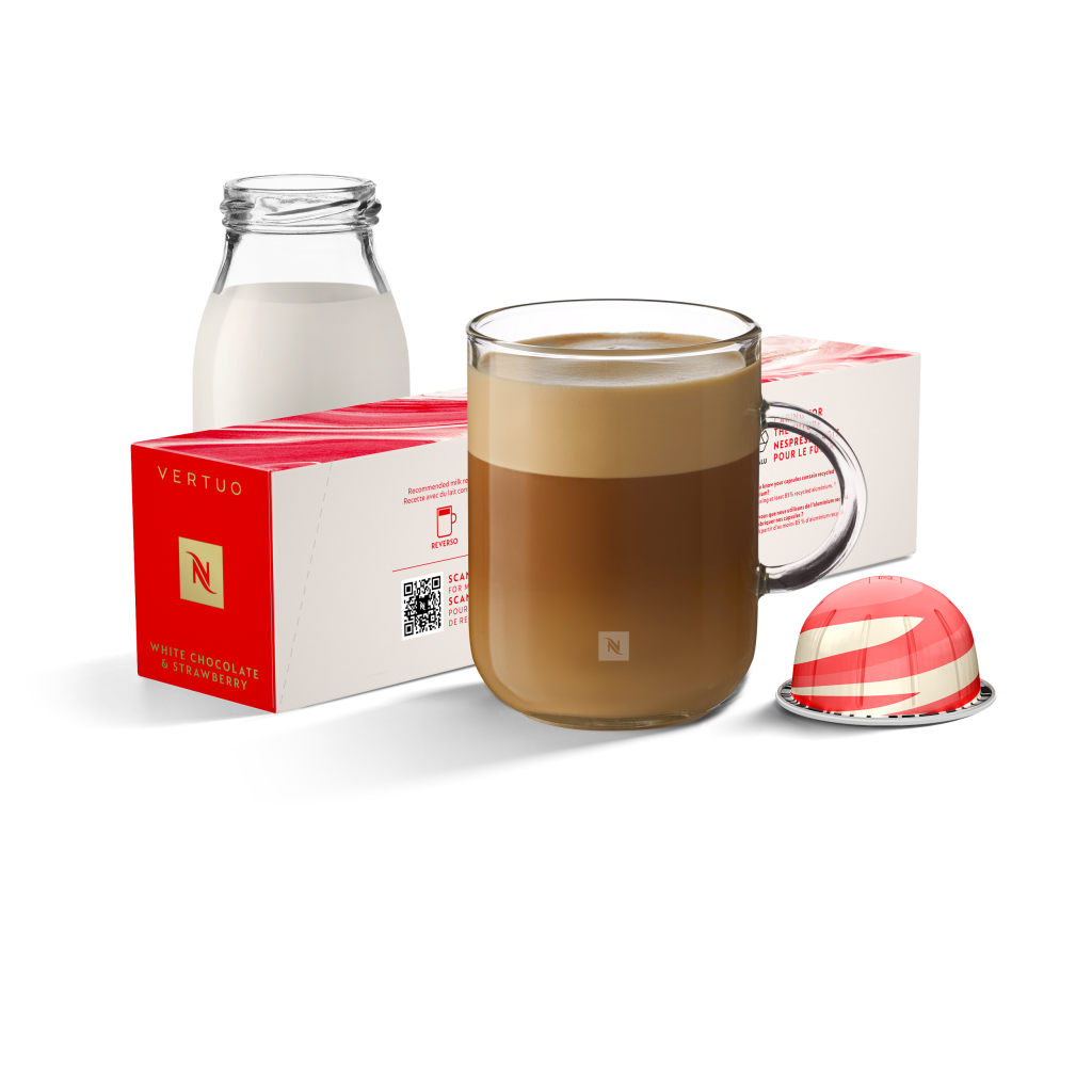 Copo de café com leite em fundo branco. Ao fundo, cápsula vermelha e branca com garrafa de leite enfeitam o ambiente.