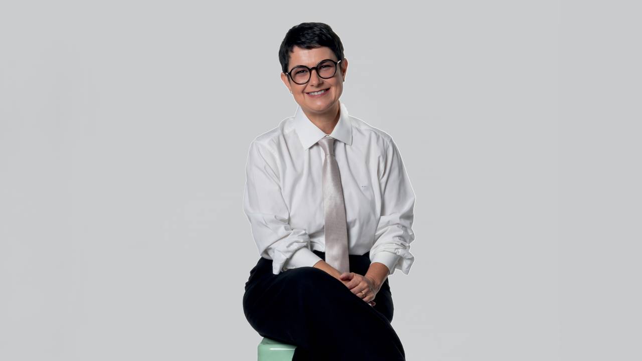 Imagem de mulher morena dos cabelos curtos, com óculos, camisa social e gravata, sorrindo.