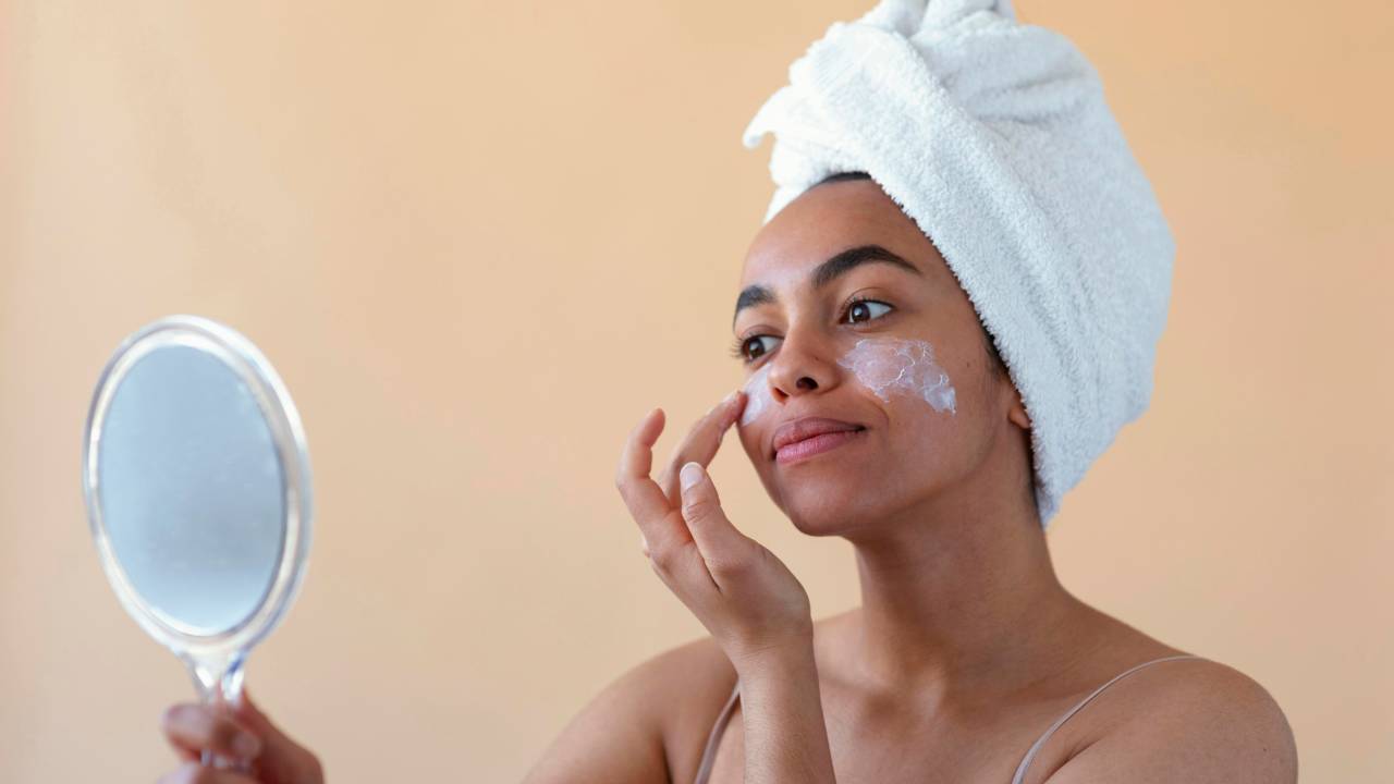 Imagem mostra mulher negra hidratando o rosto em frente ao espelho.