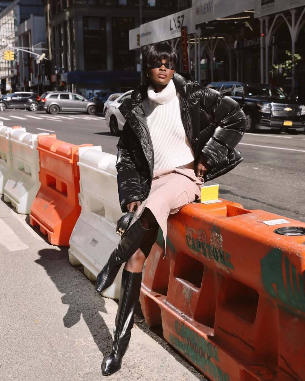 jaqueta-puffer (3) Imagem de mulher negra sentada exibindo look com jaqueta puffer oversized, saia rosa e botas de salto.