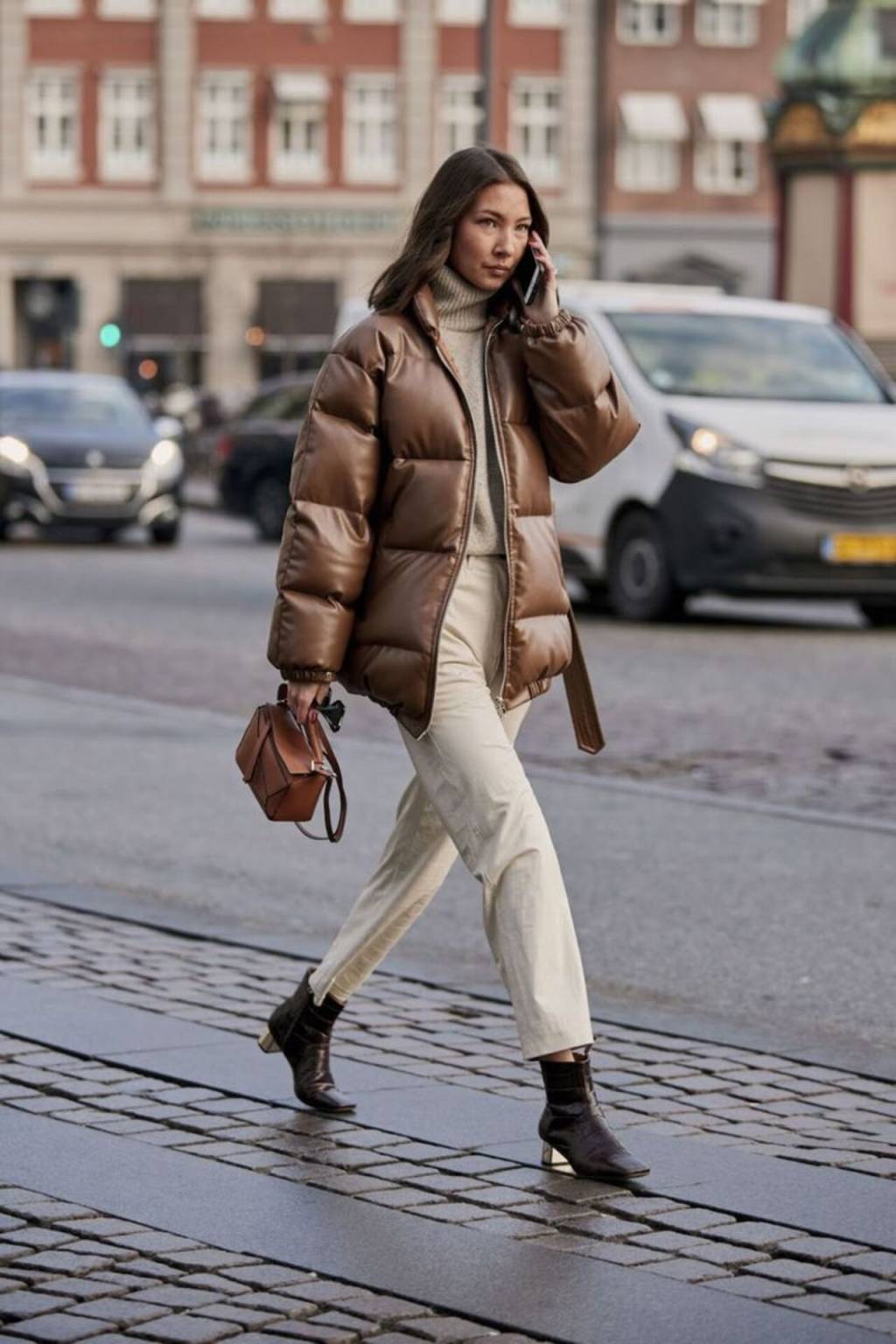 jaqueta-puffer (13) Imagem de mulher morena caminhando na rua exibindo look com jaqueta marrom, calça off white e botas de salto.