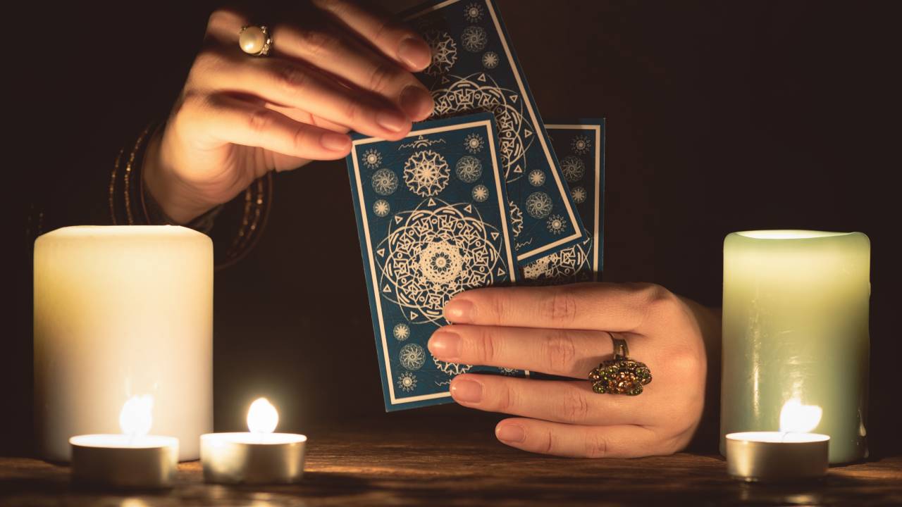 Visão das mãos de uma vidente lendo cartas de tarô na cor azul. Há duas velas amarelas: uma à esquerda e outra à direita.