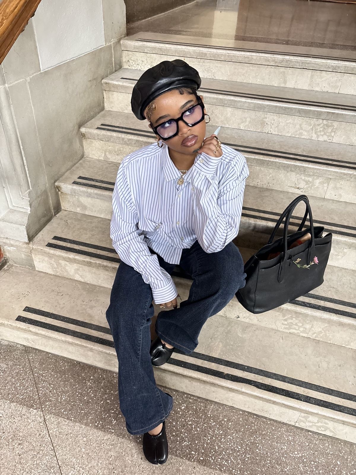 Mulher negra usando camisa listrada oversized azul, jeans escuro, boina preta e sapato tabi branco. Acessórios marcantes e look com toque retrô.