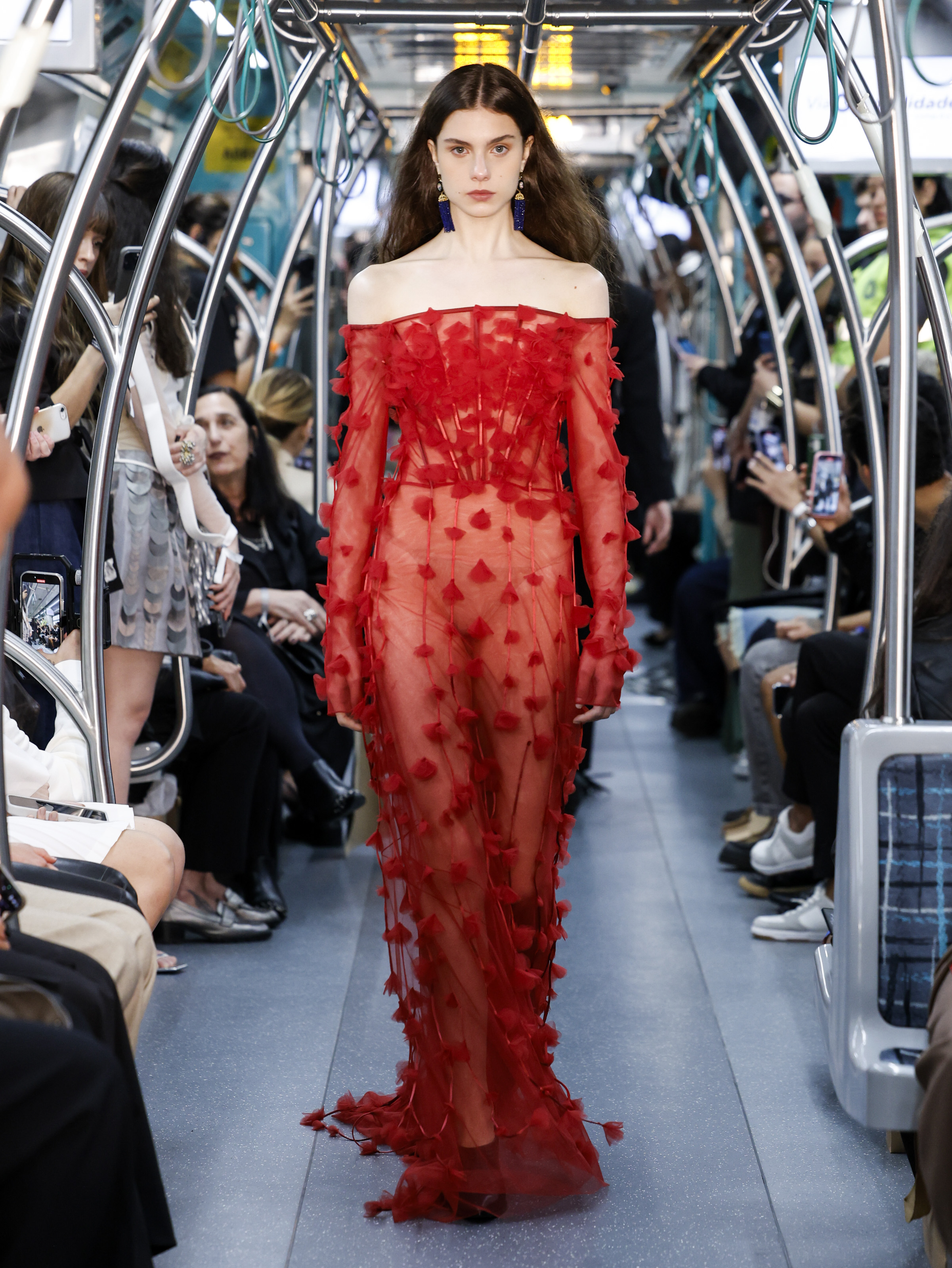 Foto de uma modelo desfilando na SPFW. Ela usa um vestido vermelho e desfila no metrô de São Paulo
