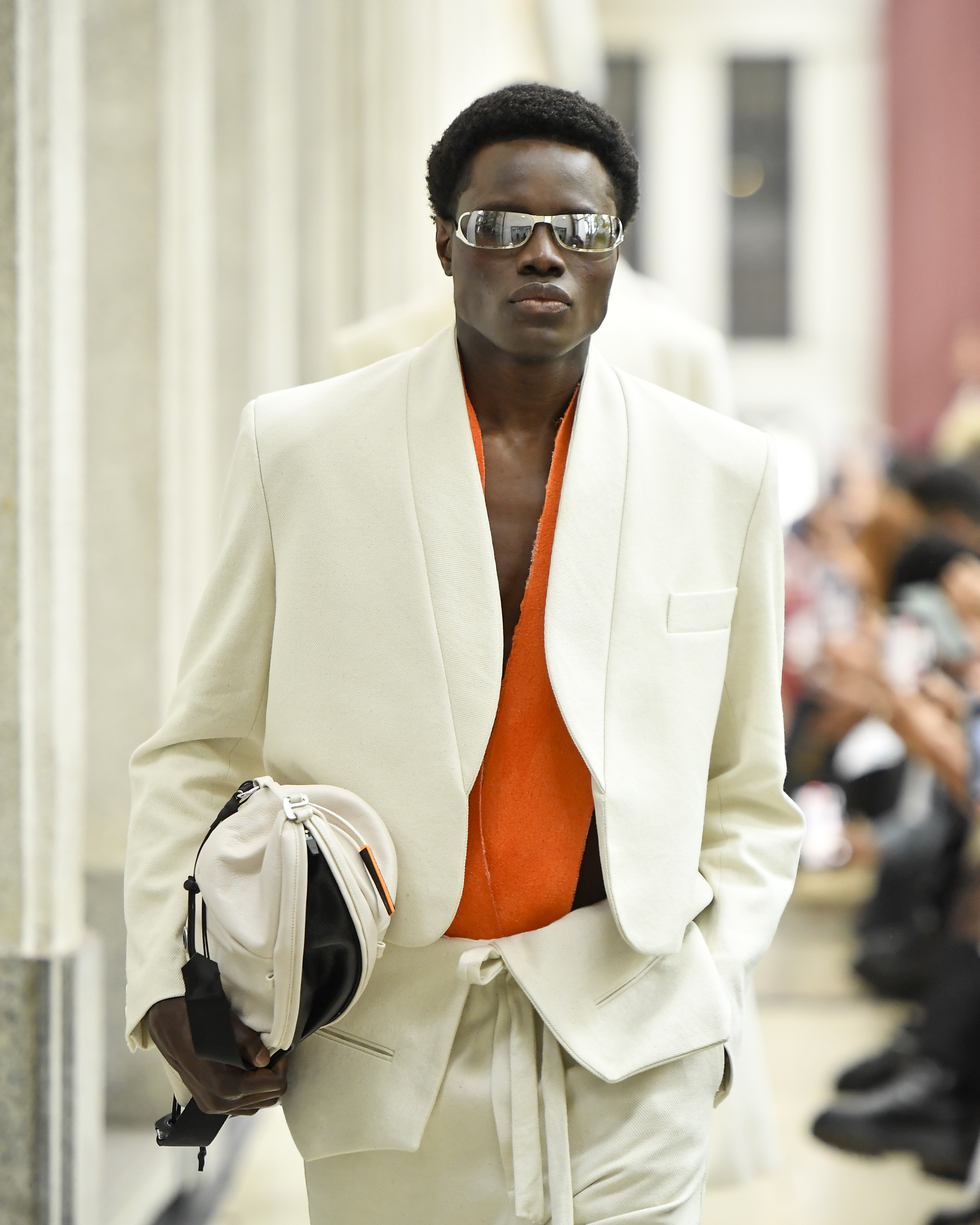 Foto de um modelo desfilando. Ele usa um blazer branco, uma blusa laranja, uma cal&ccedil;a branca e uma bolsa branca e preta